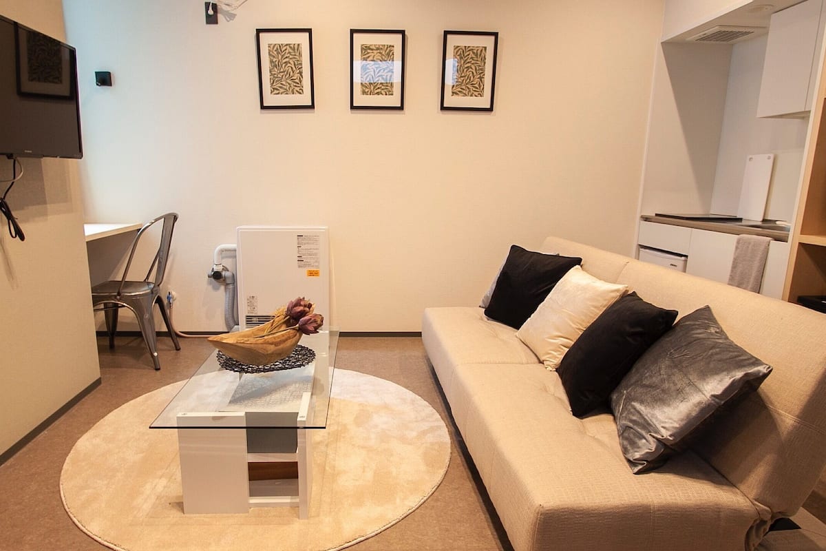 A living room with a sofa, a coffee table, a TV mounted on the wall, and a refrigerator.
