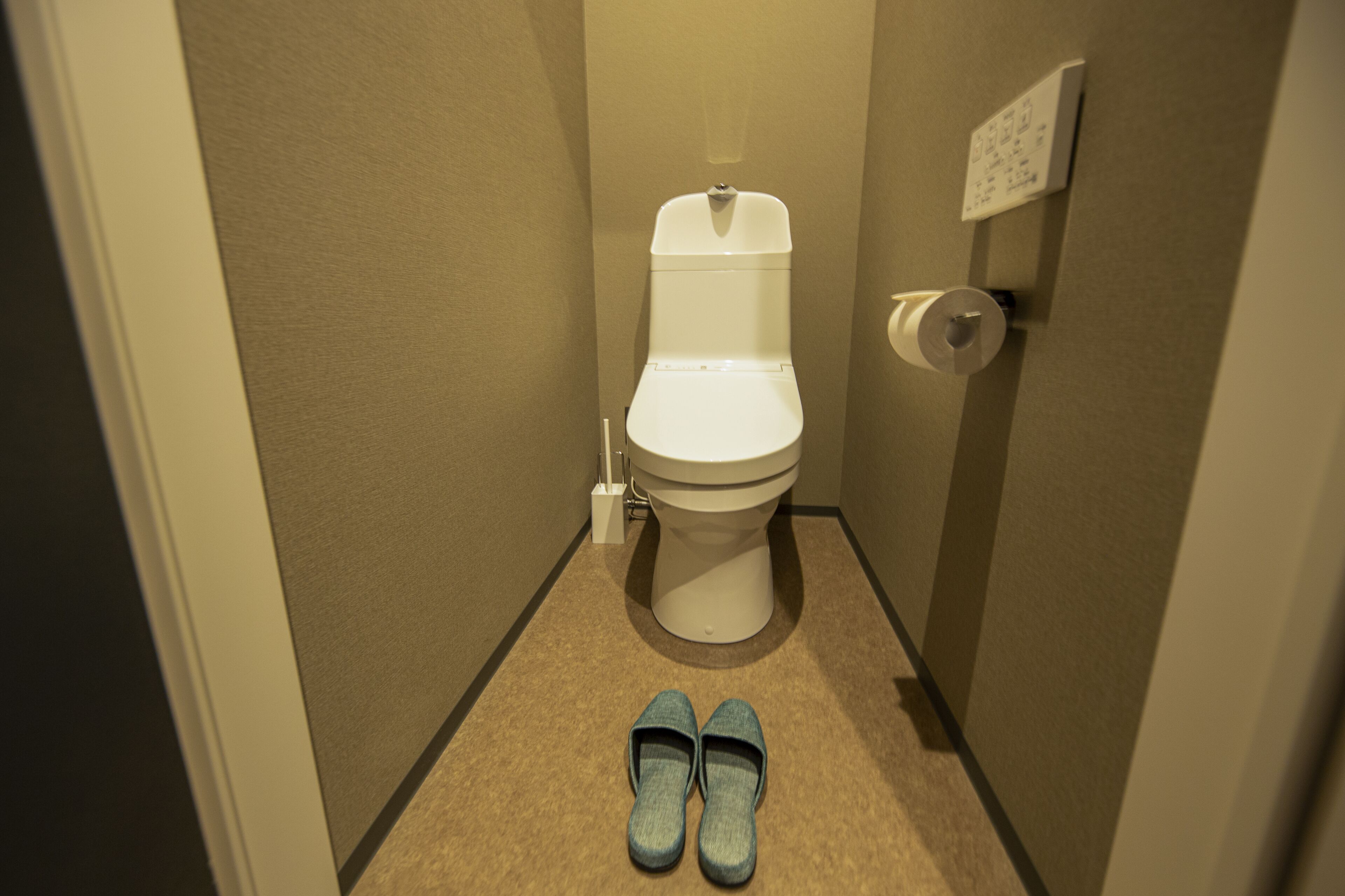 Une petite salle de bain étroite, avec des toilettes, un sèche-mains et une paire de chaussons posés sur le sol.