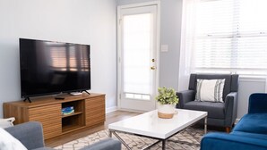 Una sala de estar moderna con una televisión de pantalla plana sobre un soporte de madera, una mesa de centro blanca, sofás grises y azules, y un ventanal con persianas.
