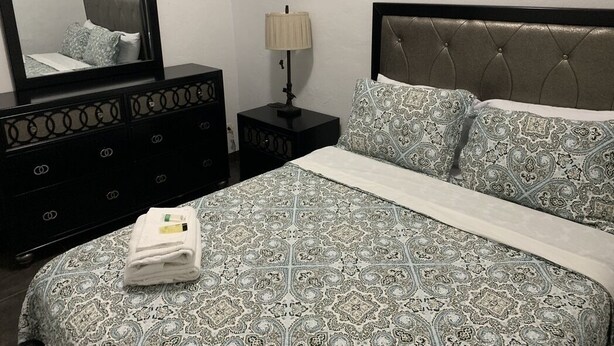 A neatly made bed with patterned bedding, a bedside lamp, a dresser with a mirror, and a headboard with tufting.
