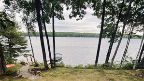 A circular seating area with Adirondack chairs around a central fire pit, overlooking a lake. - Beautiful Lakefront Retreat on Long Lake – Sleeps 11 (BRIDGTON)