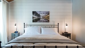 A bedroom with a bed, white walls, a mountain landscape painting, two bedside lamps, and a nightstand with a clock. - Beautiful Lakefront Retreat on Long Lake – Sleeps 11 (BRIDGTON)