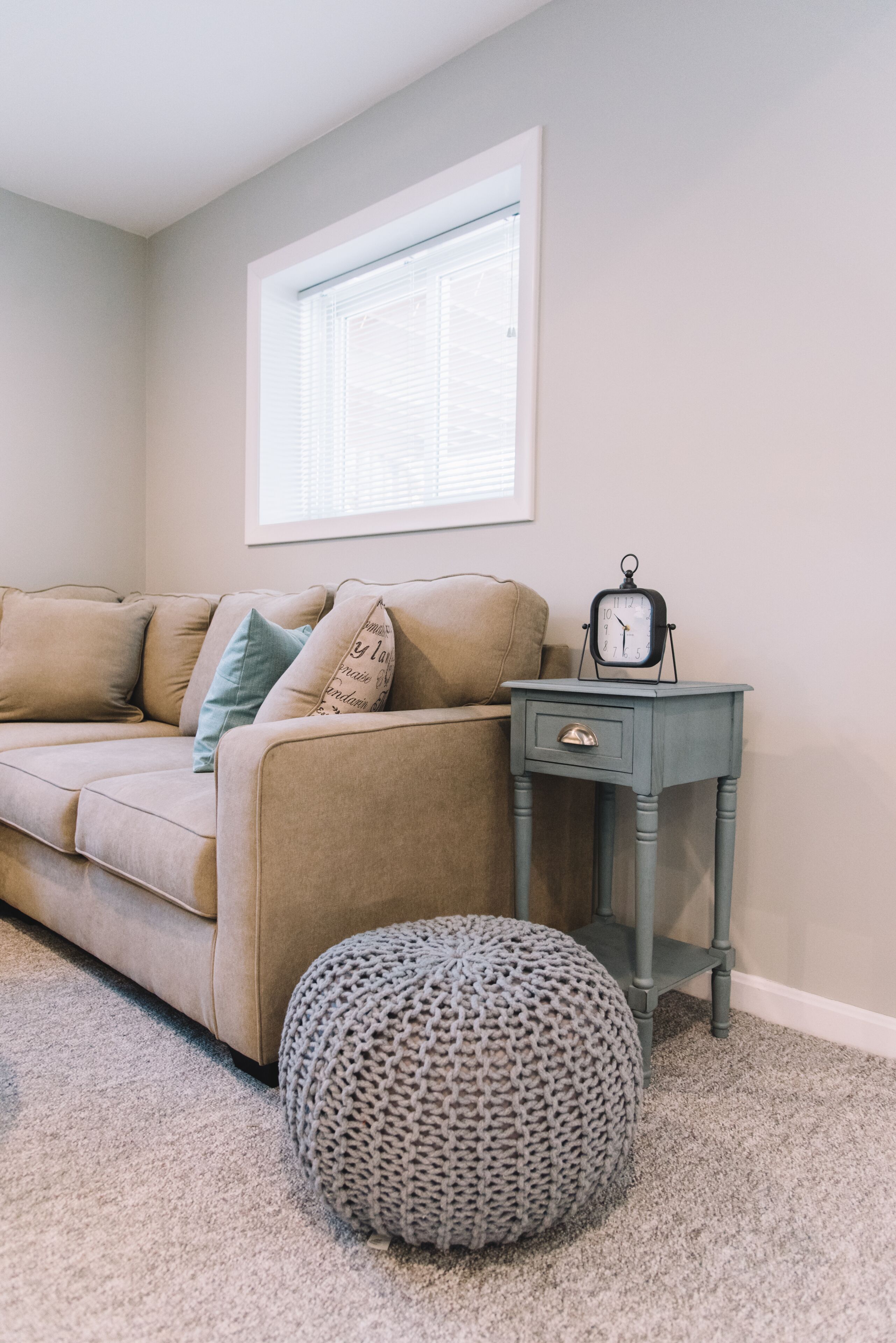 A living room with a beige sofa, a small side table with a clock, and a knitted pouf.