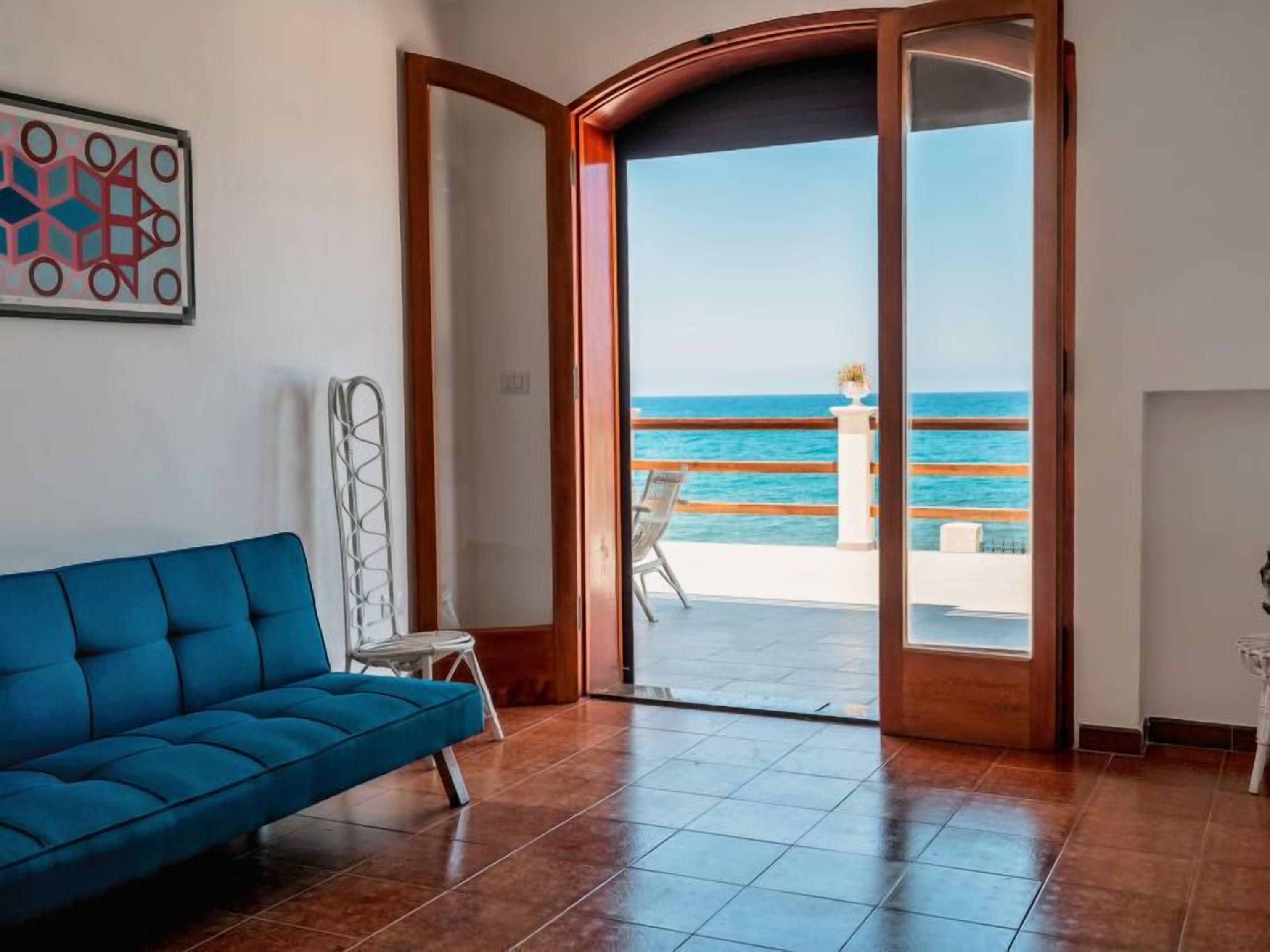 Una habitación con un sofá azul, una silla blanca y un amplio ventanal corredero que da a un balcón con vistas al mar.