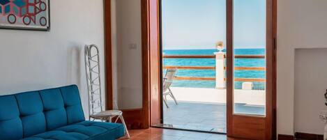 Una habitación con un sofá azul, una silla blanca y un amplio ventanal corredero que da a un balcón con vistas al mar.