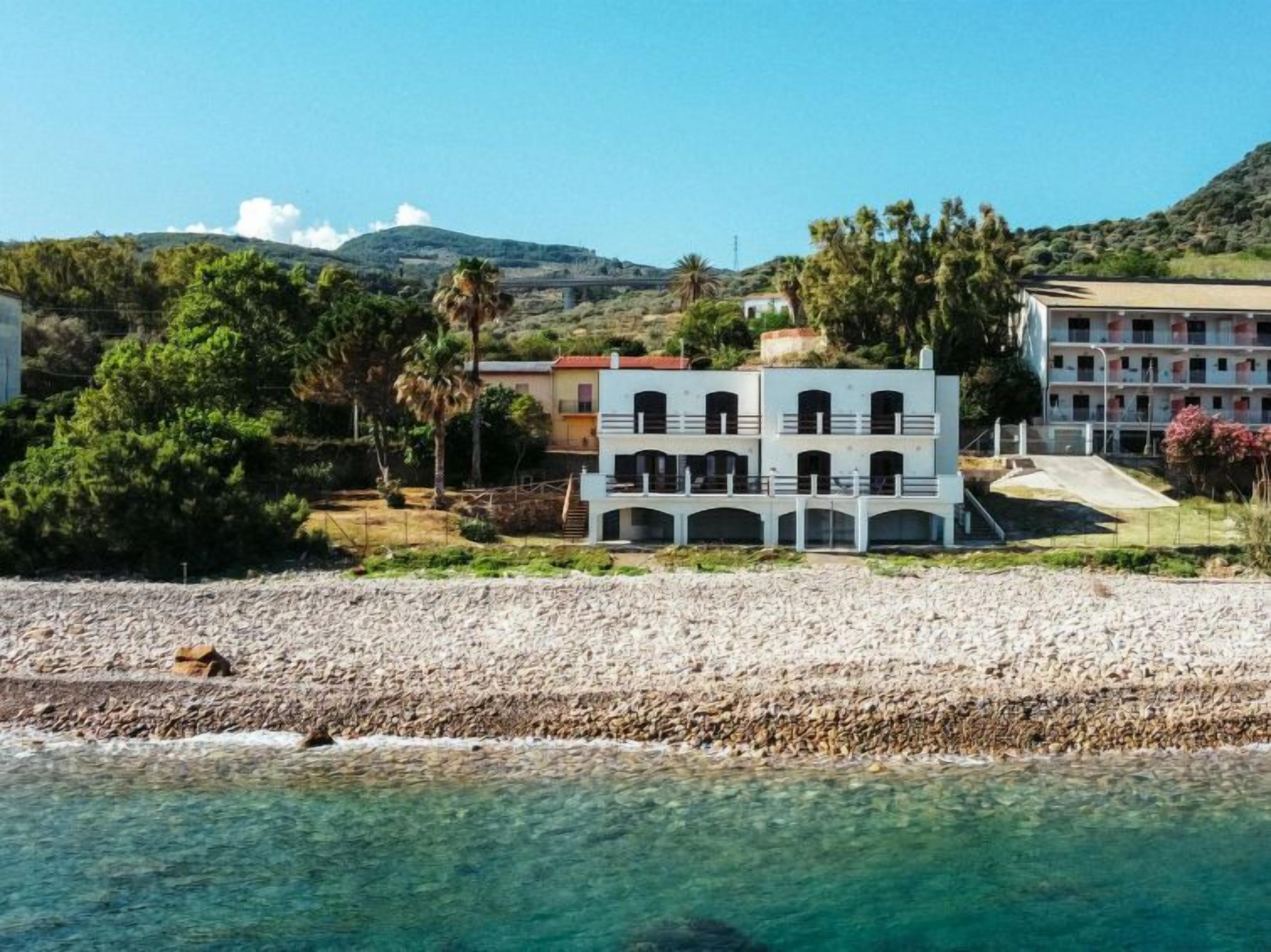 Un bâtiment situé sur la côte, doté de nombreux balcons, donnant sur une plage de galets et bordant une eau d’un bleu limpide.