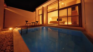 Un área de piscina moderna al aire libre, con vista despejada al interior a través de amplios ventanales, una escalera que conduce a un balcón en un segundo piso y una pequeña zona de estar exterior.