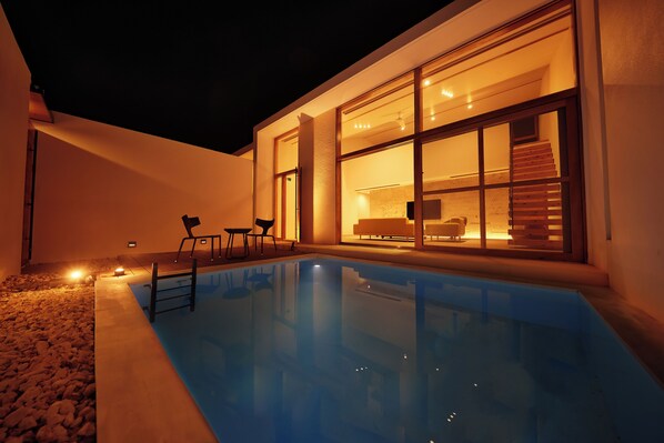 A modern outdoor pool area with a clear view of the interior through large windows, a set of stairs leading to a second-floor balcony, and a small outdoor seating area.