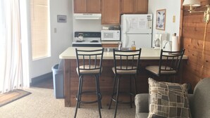 A compact kitchen with wooden cabinets, a white countertop, a stove, a microwave, and a refrigerator. There are three bar stools and a small island. - Hidden Valley - Walk to Slopes and Pool - 2 BR with Fireplace + WIFI (Hidden Valley)