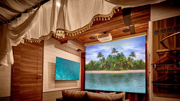 A room with a large flat-screen TV displaying a tropical beach scene, a wooden ceiling with a hanging light fixture, and a brown leather sofa. - Petit Bali Ikebukuro - Adults Only (Tokyo)
