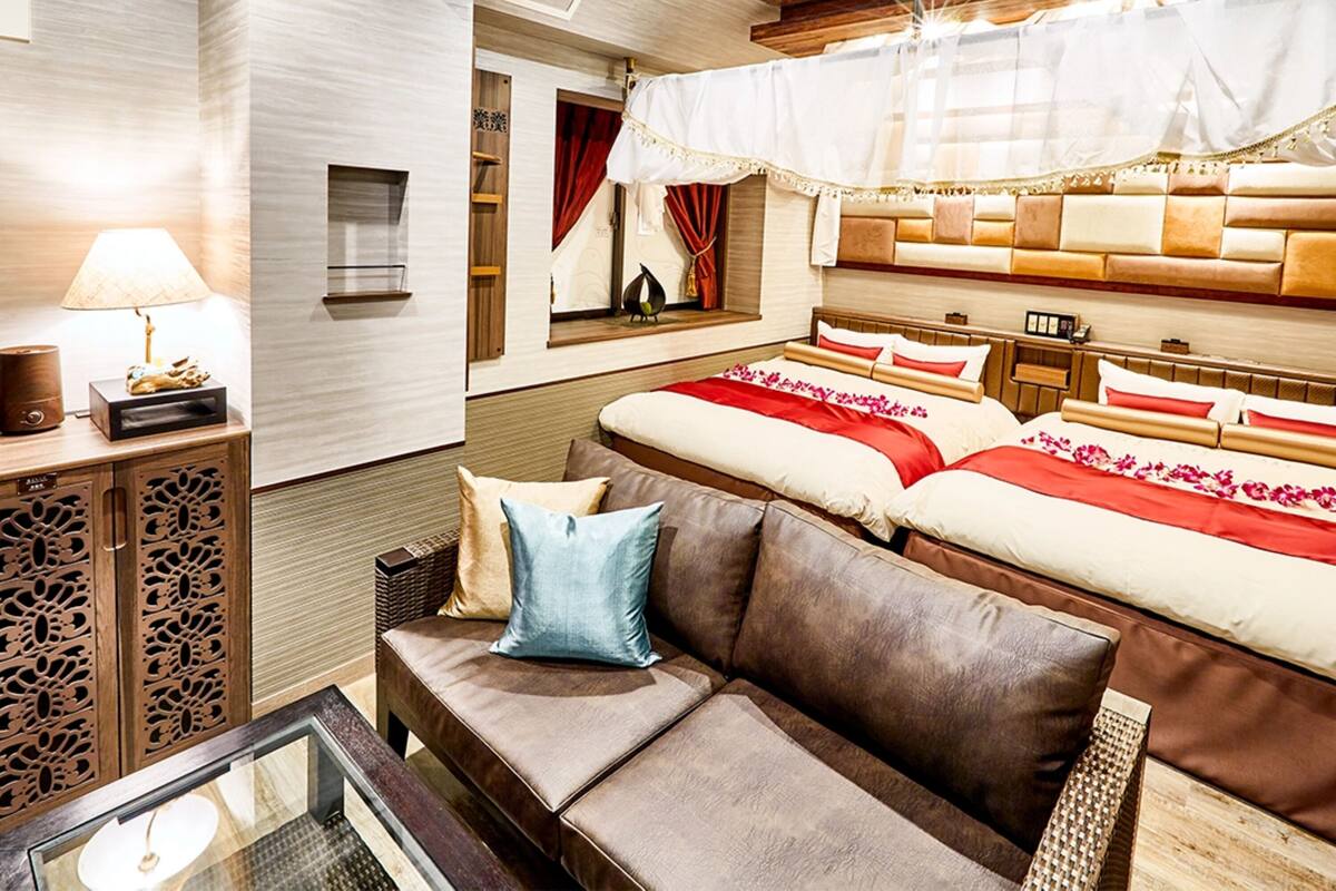A hotel room with a wooden ceiling, a large bed with red and white bedding, a brown sofa, and a small table with a lamp.
