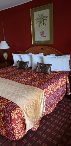 A hotel room with a large bed, red walls, a framed picture of a plant, and a lamp on a nightstand. - Palms Inn (Dania Beach)