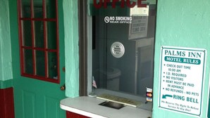 A motel office with a green door, a window with a 'Motel Office' sign, a 'No Smoking' sign, and a 'Palms Inn Motel Rules' sign listing check-out time, ID requirement, no visitors, advance payment, and no refunds or pets. - Palms Inn (Dania Beach)