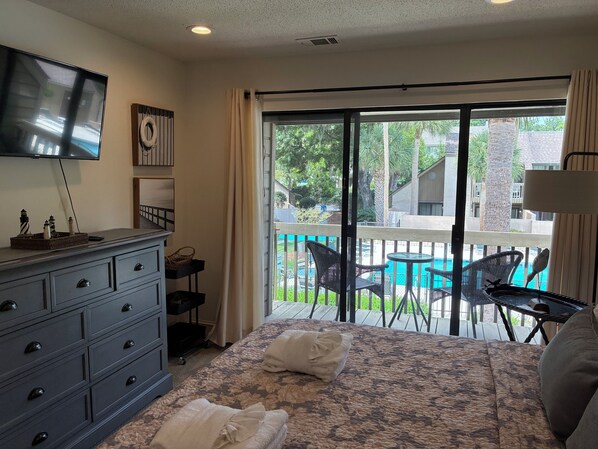 A bedroom with a bed, a dresser, a television, a window with a view of a pool and outdoor area.