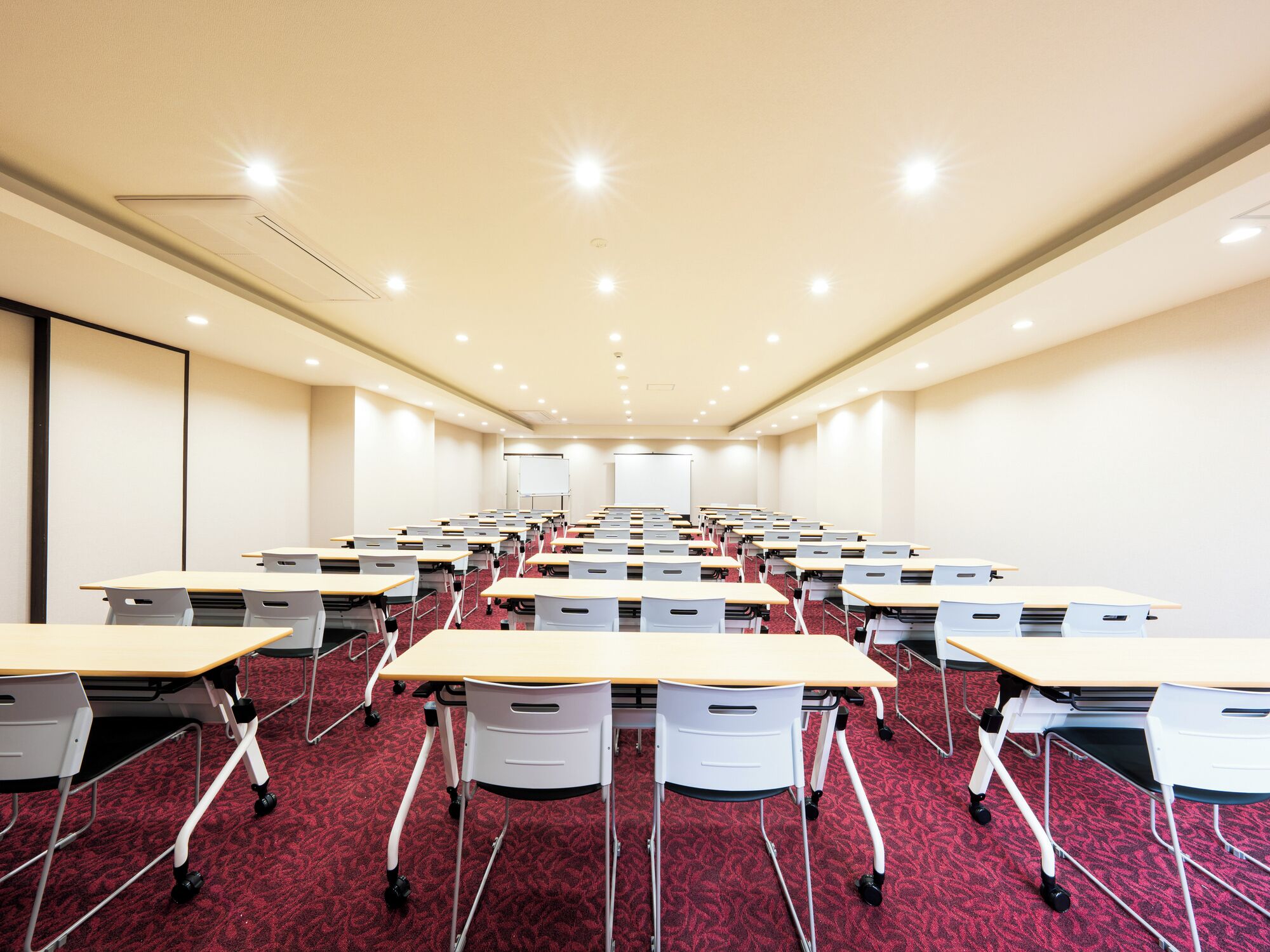 Uma sala de conferências com fileiras de mesas e cadeiras, um quadro branco e uma tela de projeção.