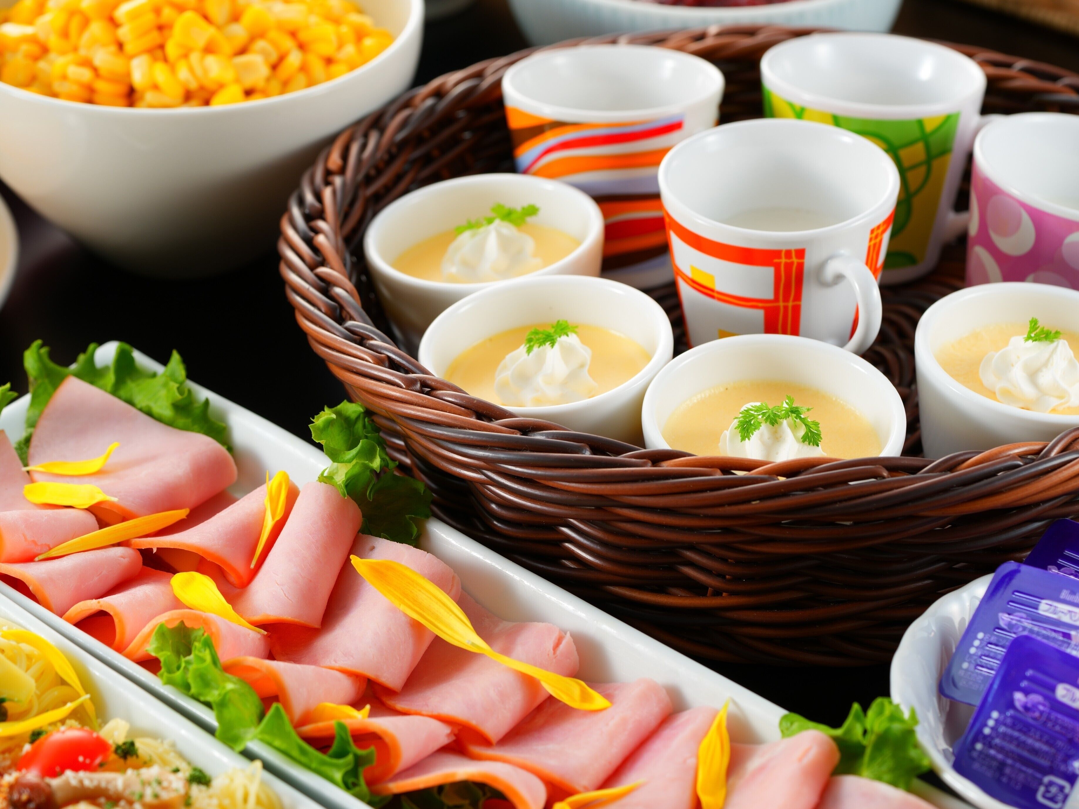 A buffet with sliced meats, bowls of soup, and colorful cups.