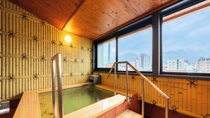Perinteinen japanilainen onsen-kylpylä, jossa on puuseinät, puinen kylpyallas ja suuret ikkunat, joista avautuu näkymä kaupungin siluettiin.