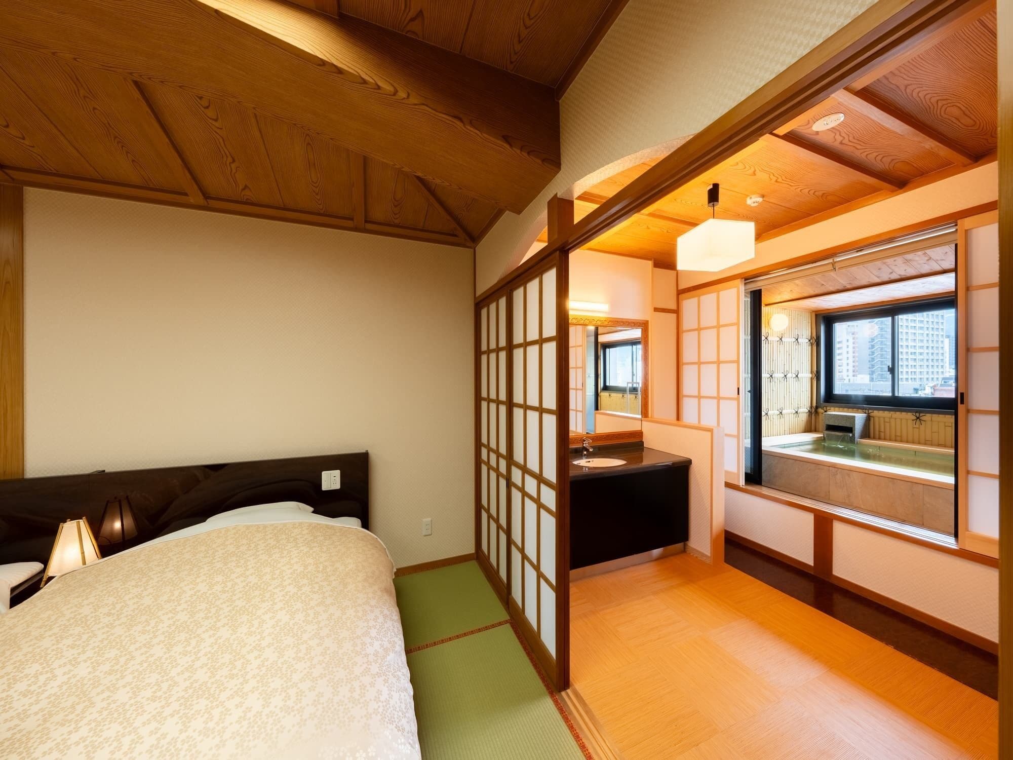 A traditional Japanese-style room with tatami flooring, a bed with a wooden headboard, a washbasin with a mirror, and a window with a view of the city.