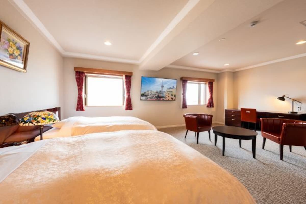 A hotel room with two beds, a desk, a chair, a table, and a painting on the wall.