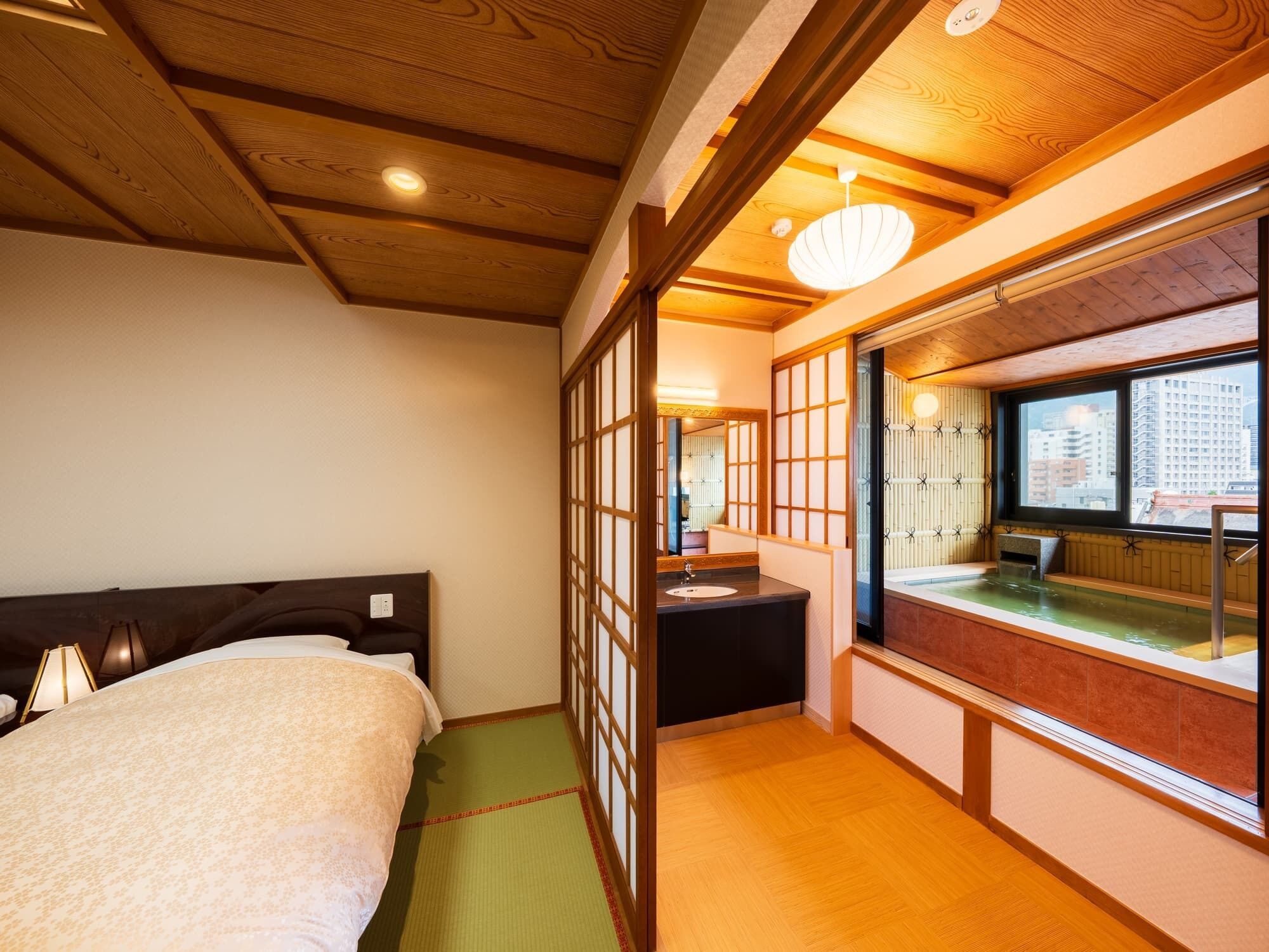 Una habitación japonesa tradicional con suelo de tatami, una cama, un lavamanos y vistas de un paisaje urbano a través de un ventanal grande.
