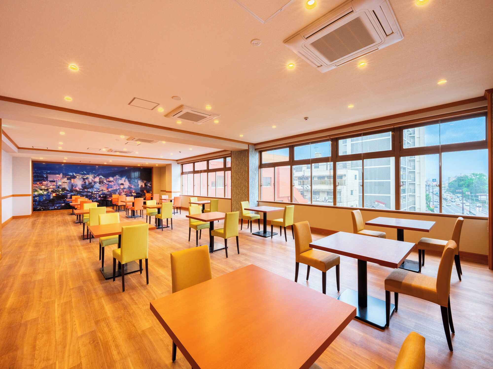 A spacious dining area with multiple tables and chairs, large windows offering a city view, and a mural depicting a cityscape on the far wall.