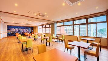 A spacious dining area with multiple tables and chairs, large windows offering a city view, and a mural depicting a cityscape on the far wall.