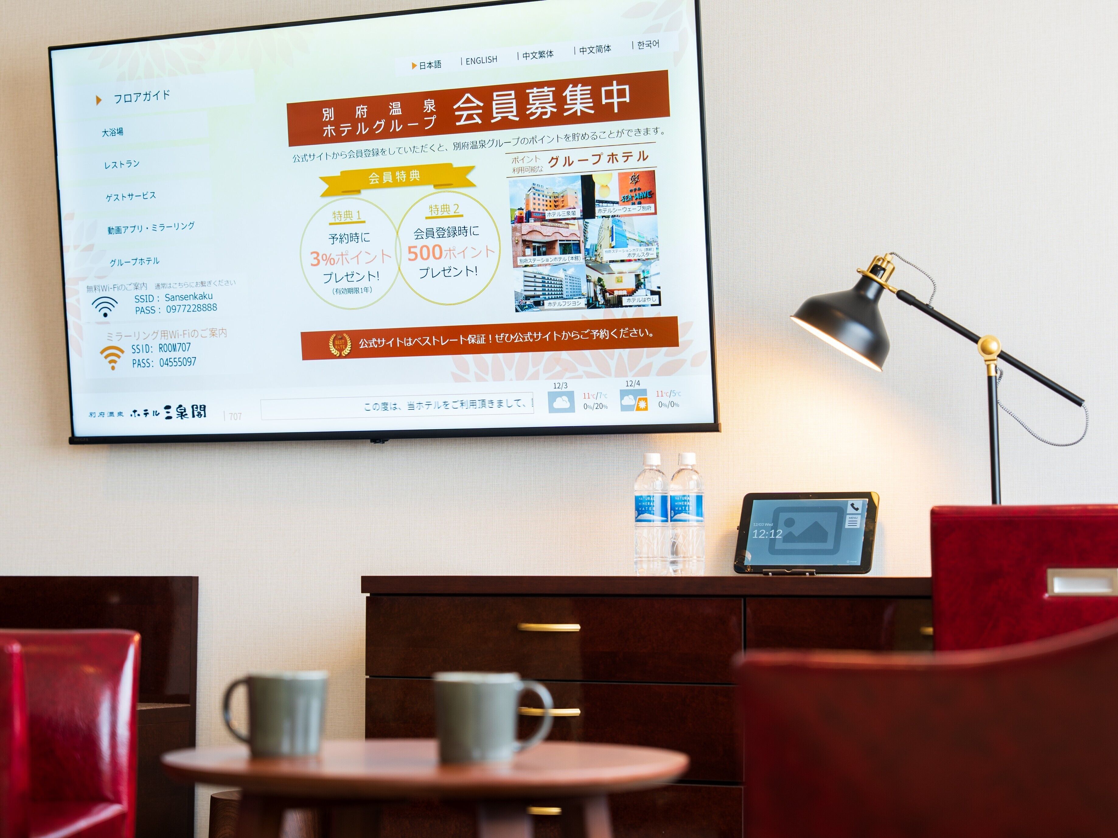 Een hotelkamer met een groot scherm waarop de hotelwebsite staat, een bureau met een laptop, een lamp en twee waterflessen op het nachtkastje.