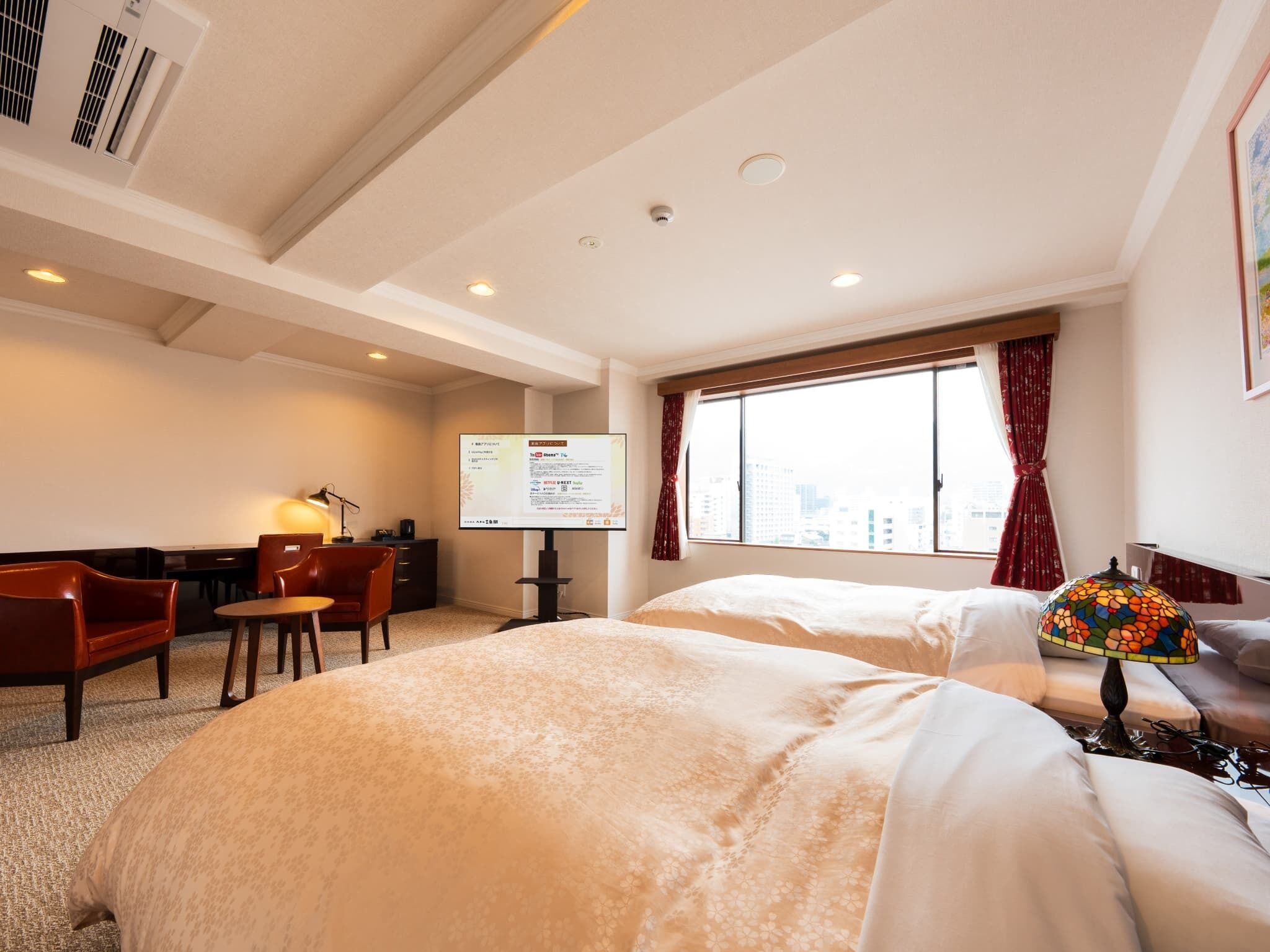 A hotel room with two beds, a desk with a chair, a TV, and a window with curtains.