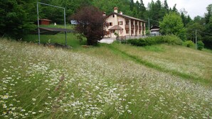 Un bâtiment à deux étages doté d’un balcon, entouré d’un champ verdoyant parsemé de fleurs blanches, avec des montagnes en toile de fond.