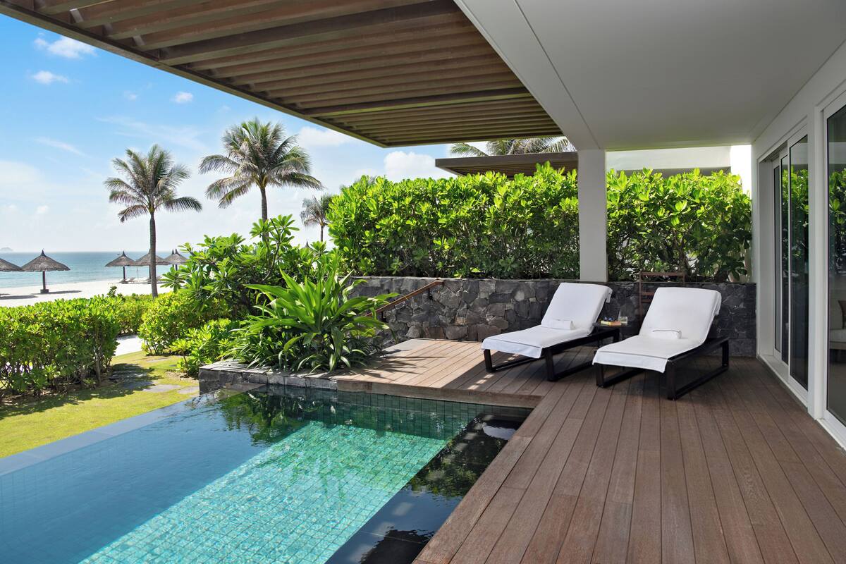 An infinity pool with a wooden deck, two white lounge chairs, and a view of the beach and ocean.