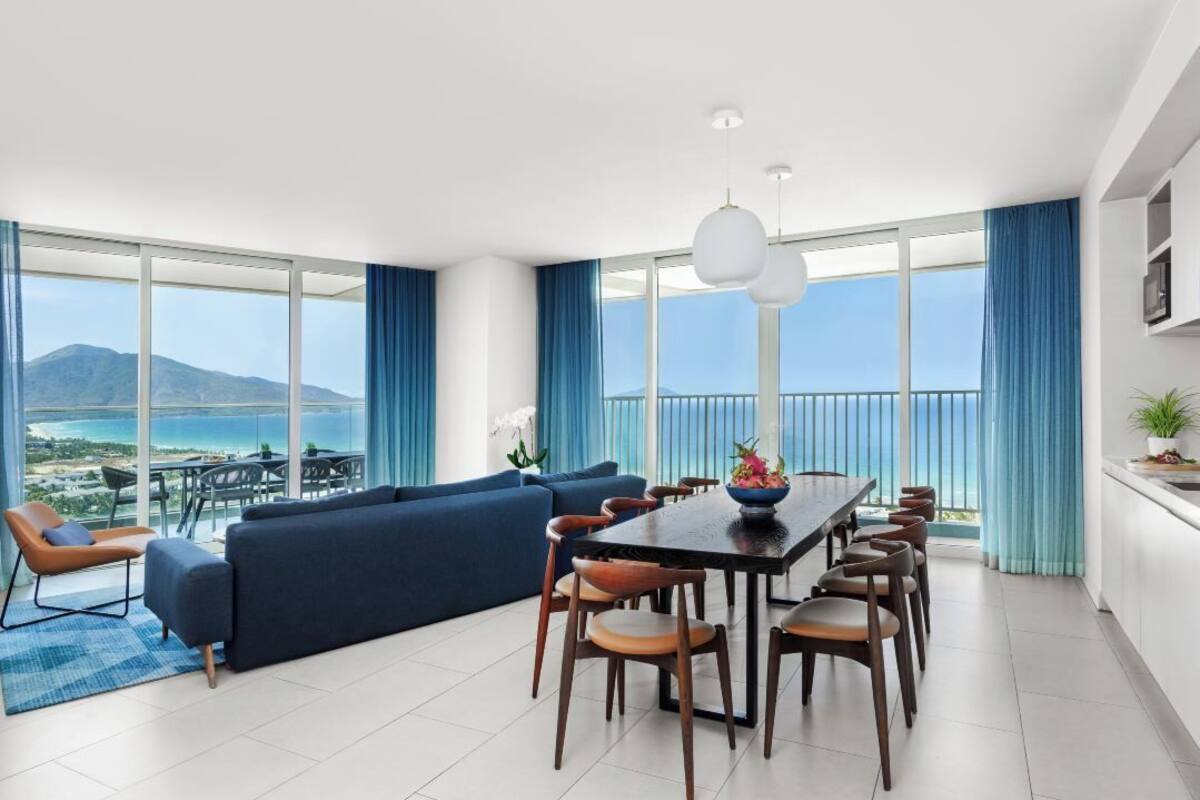 A modern living room with a large sofa, dining table, and chairs, large windows with a view of the sea and mountains, and a balcony with a view.