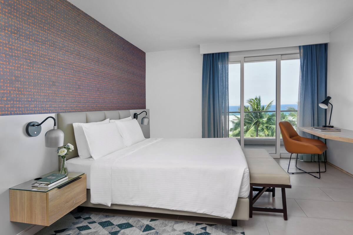 A modern hotel room with a large bed, a desk, and a view of the ocean through the window.