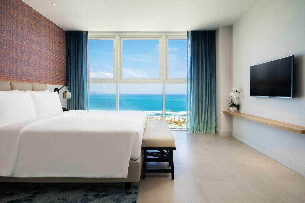 A modern hotel room with a large bed, a TV mounted on the wall, and a view of the ocean.