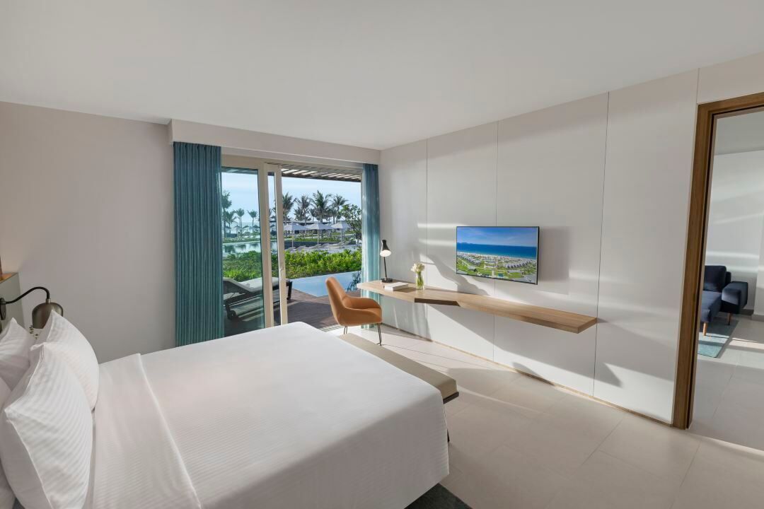 Een moderne hotelkamer met een groot bed, een bureau met stoel, een televisie en uitzicht op een zwembad en palmbomen.