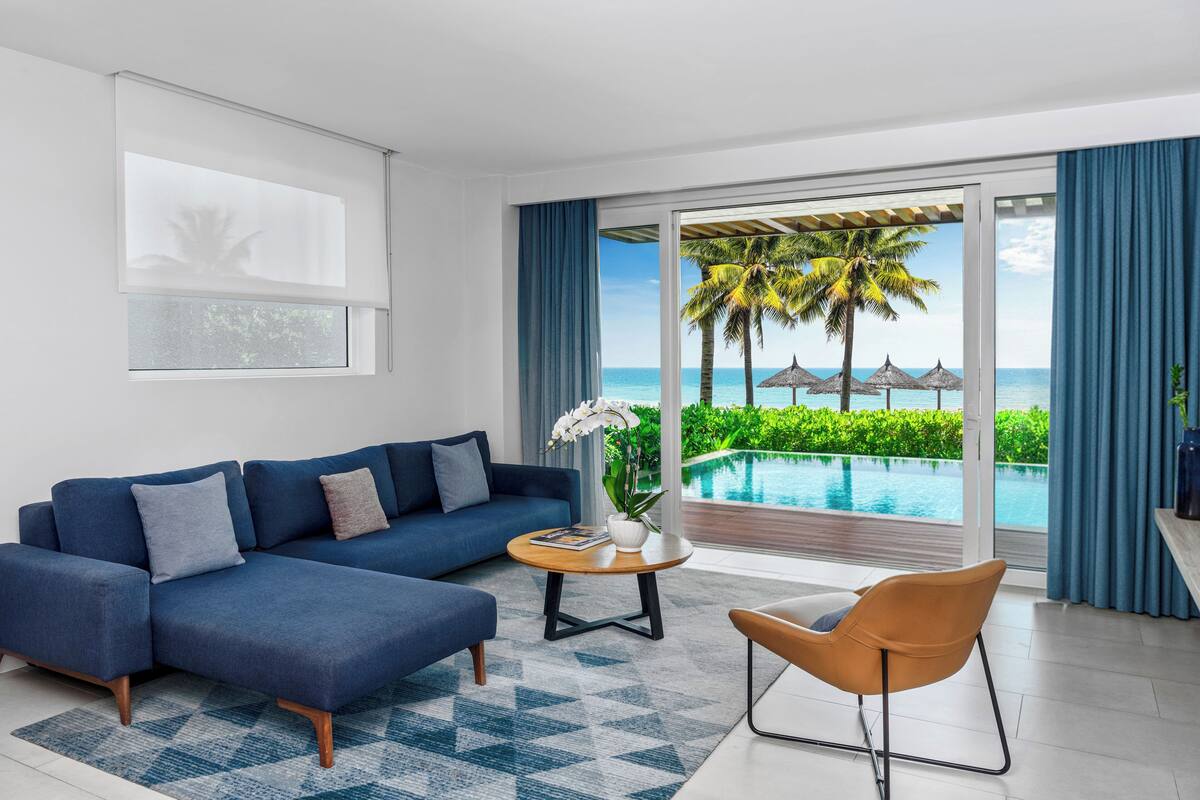 A modern living room with a sofa, a coffee table, and a chair. A large window offers a view of a pool and palm trees.