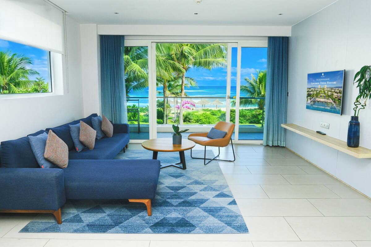 A modern living room with a blue sofa, a round coffee table, and a chair. There is a large window with a view of palm trees and the ocean.