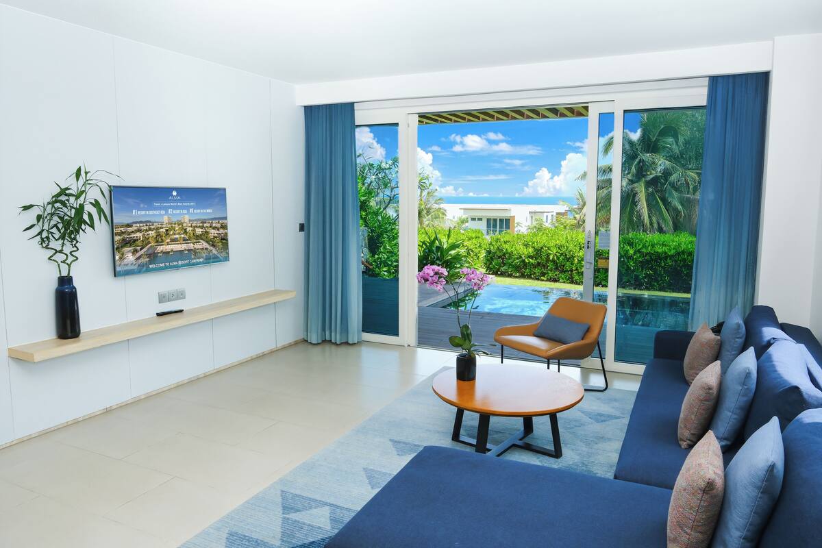 A modern living room with a sofa, a coffee table, and a chair. There is a large window with a view of a pool and greenery.