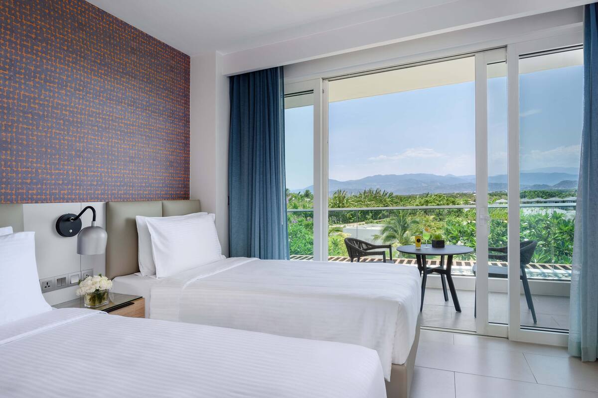 A hotel room with two beds, a nightstand with a lamp, a small table with a chair, and a balcony with a view of greenery and mountains.