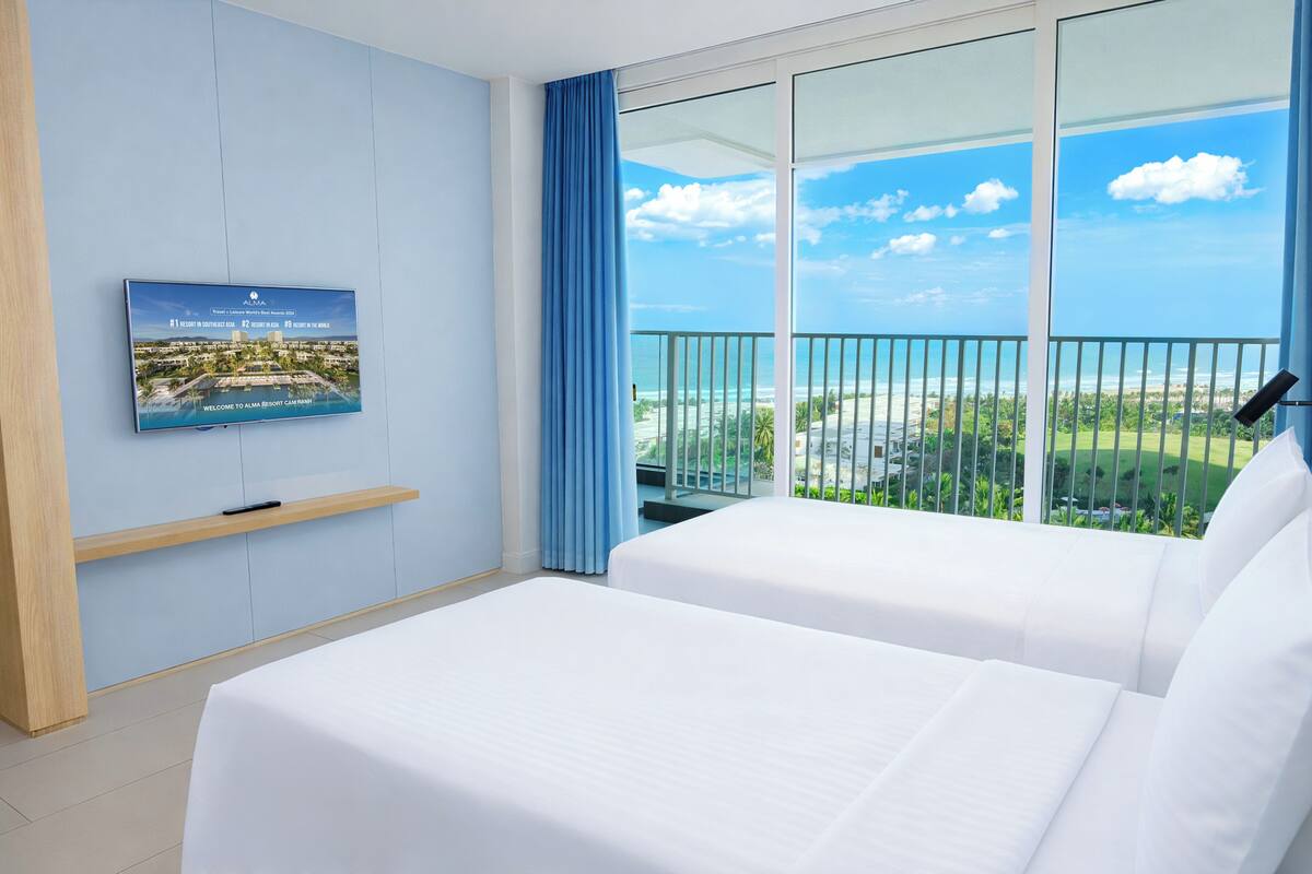 A hotel room with two beds, a TV on the wall, and a balcony with a view of the ocean.