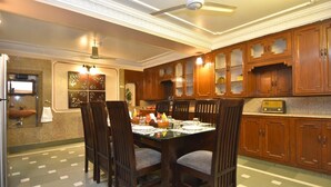 Uma sala de jantar com uma mesa posta para quatro pessoas, rodeada por cadeiras de madeira. Um ventilador de teto é visível acima da mesa, e existem armários de madeira com portas de vidro à direita.