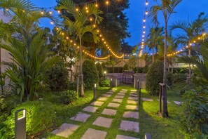 A garden pathway with string lights, palm trees, and a barbecue area.