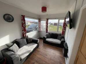 A living room with a sofa, a smaller chair, a flat-screen TV, and a clock on the wall. - Captivating 2-bed Chalet (Bridlington)