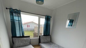 A room with two single beds, a window with curtains, a small bedside table, and a mirror on the wall. - Captivating 2-bed Chalet (Bridlington)