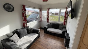 A living room with a sofa and a chair, a clock on the wall, and a TV mounted on the wall. - Captivating 2-bed Chalet (Bridlington)