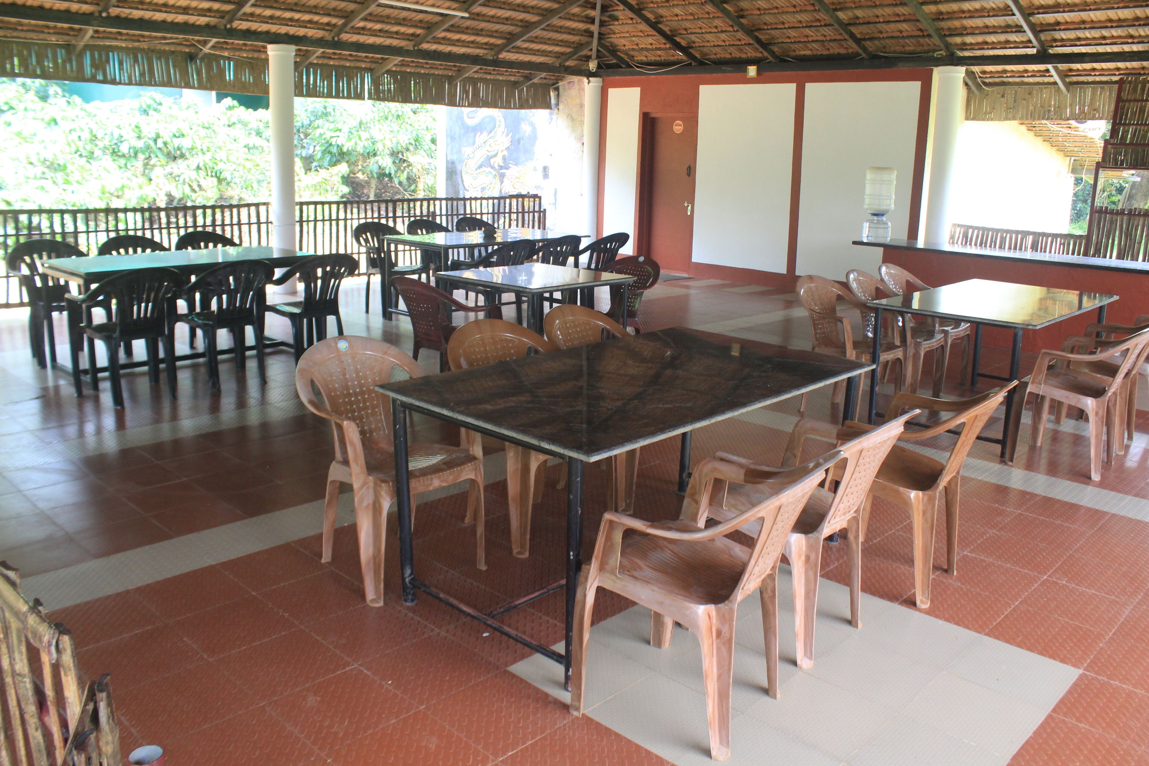 Un espace de restauration abrité, doté de plusieurs tables et chaises, d’un sol carrelé et offrant une vue sur la verdure environnante.