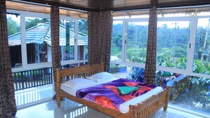 A wooden bed with a colorful blanket in a room with large windows and curtains. - Muccole Stay (Madikeri)