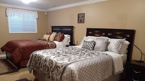 A bedroom with two beds, a large bed with a patterned bedspread and pillows, and a smaller bed with a red blanket. There is a window with blinds, a nightstand with a lamp, and a painting on the wall. - Spacious Two-Bedroom Suite in Salt Lake Valley (West Jordan)