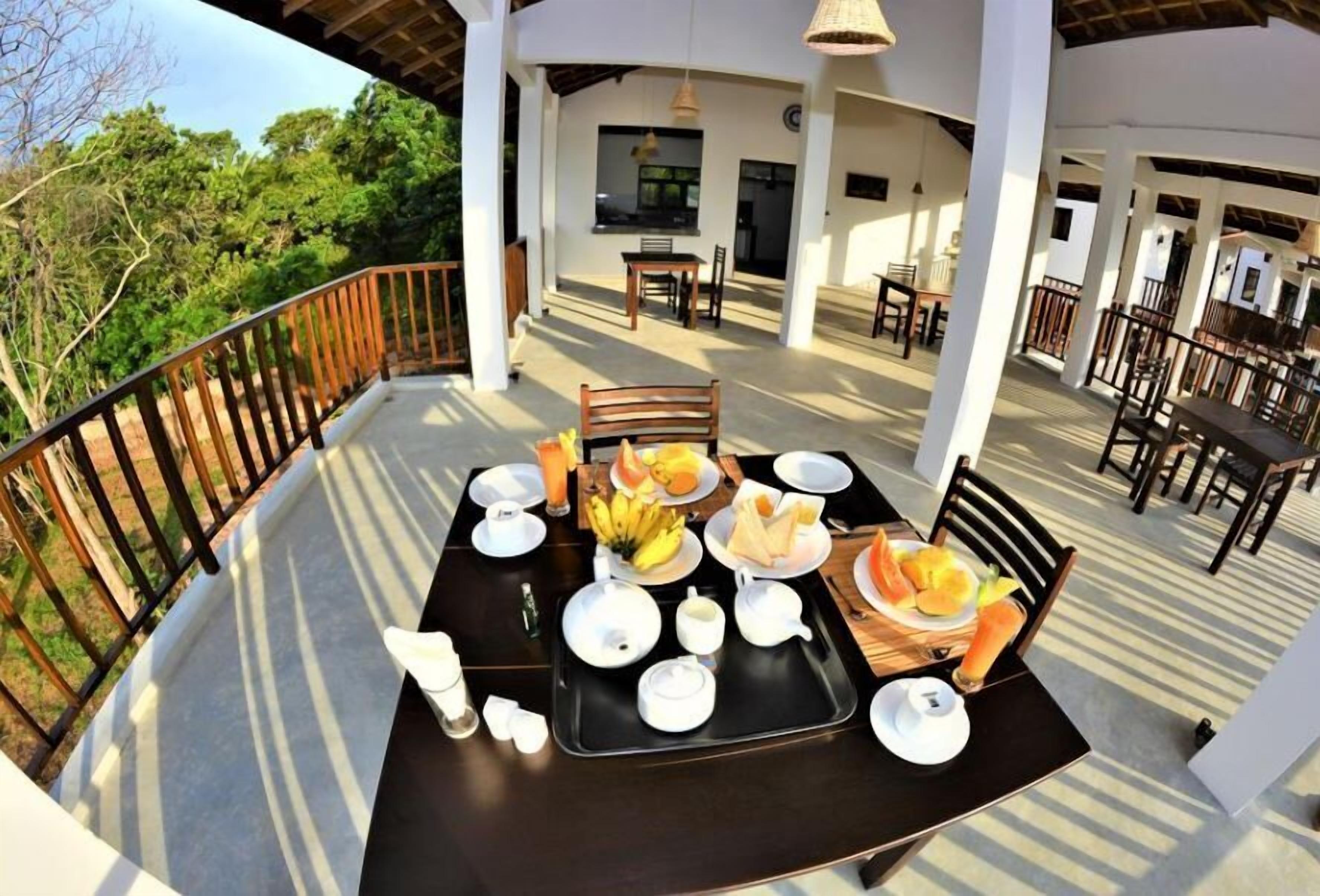 A breakfast setup on a balcony with a view of trees, a table with plates, bowls, and a fruit basket.