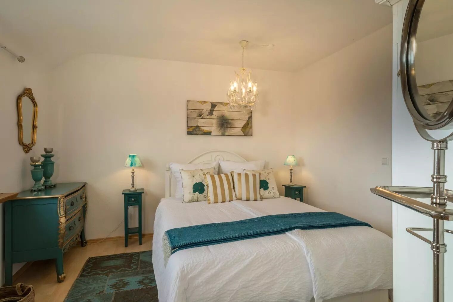A bedroom with a bed, two bedside tables, a chandelier, a dresser, a mirror, and a wall-mounted artwork.