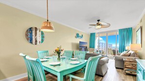 A dining area with turquoise chairs and a table, a living room with a sofa and a TV, and a balcony with a view.